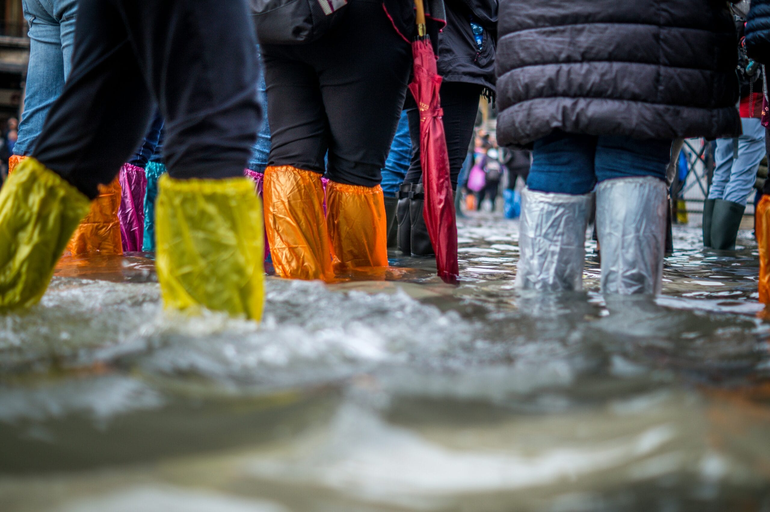 Colorful waterproof boots wade through shallow floodwaters, with blurred figures in the background.