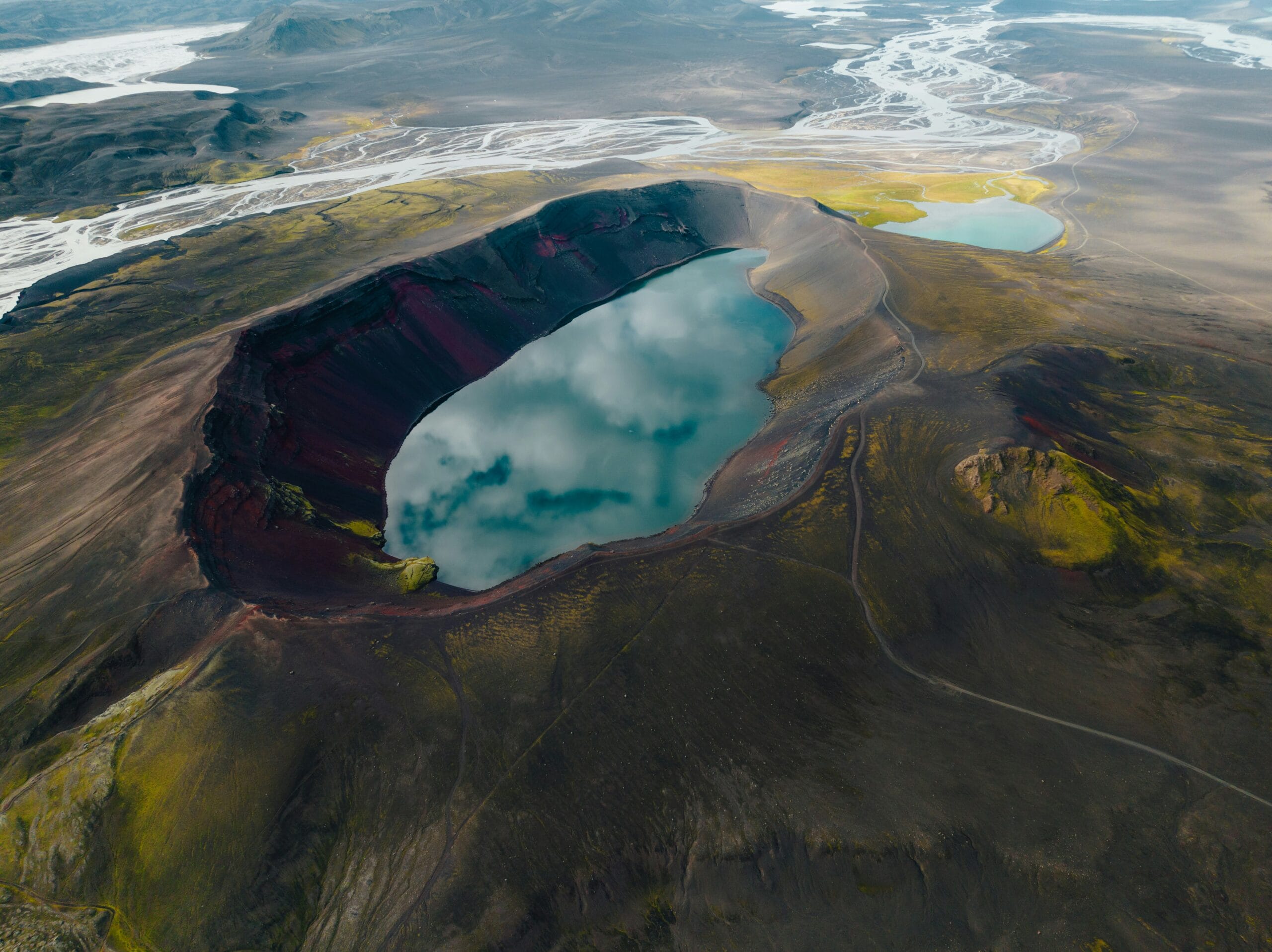 Aerial view of a volcanic crater filled with reflective water, surrounded by rugged terrain and distant rivers.