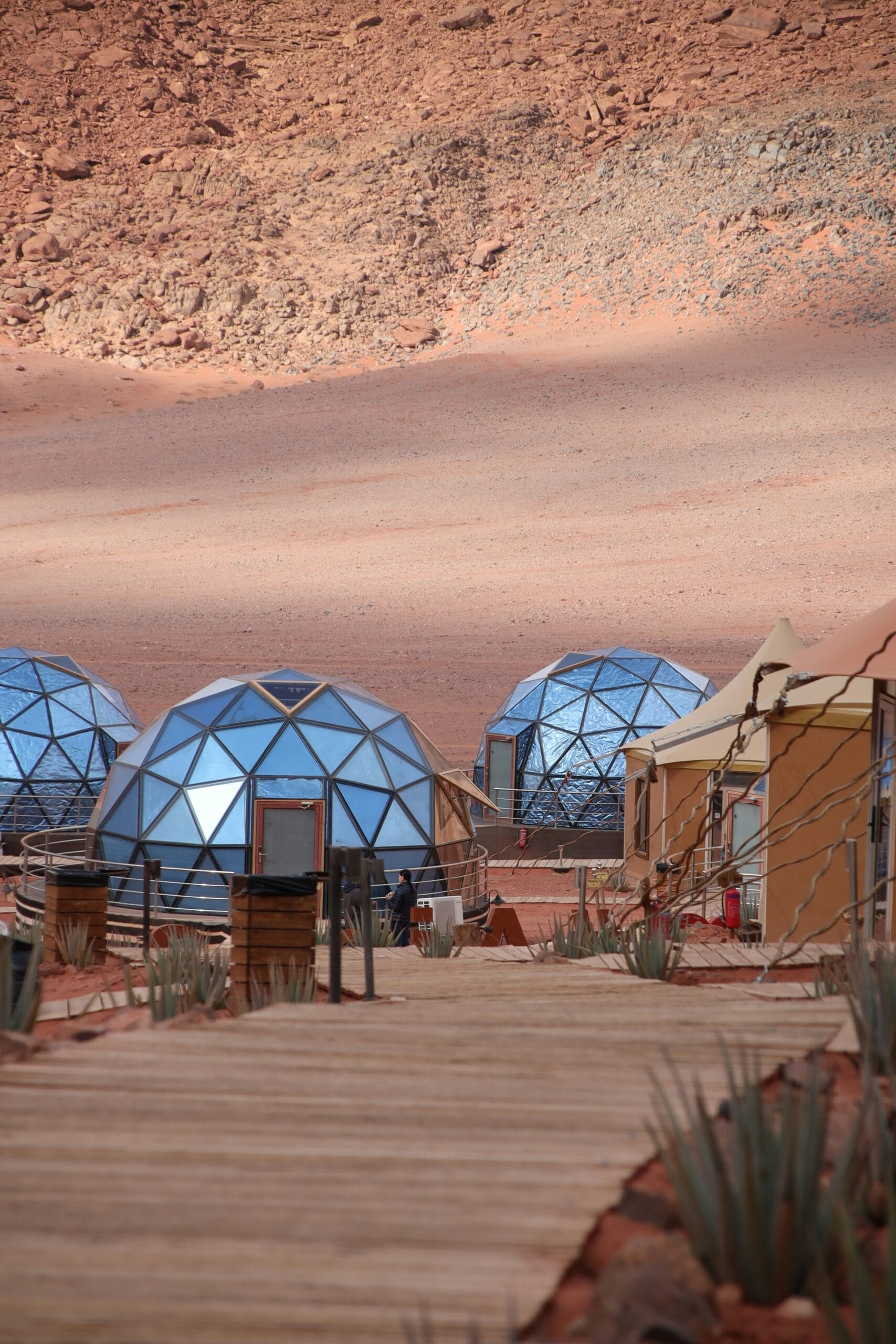 Geodesic domes and tents set against a rocky desert landscape, connected by wooden pathways and surrounded by sparse vegetation.