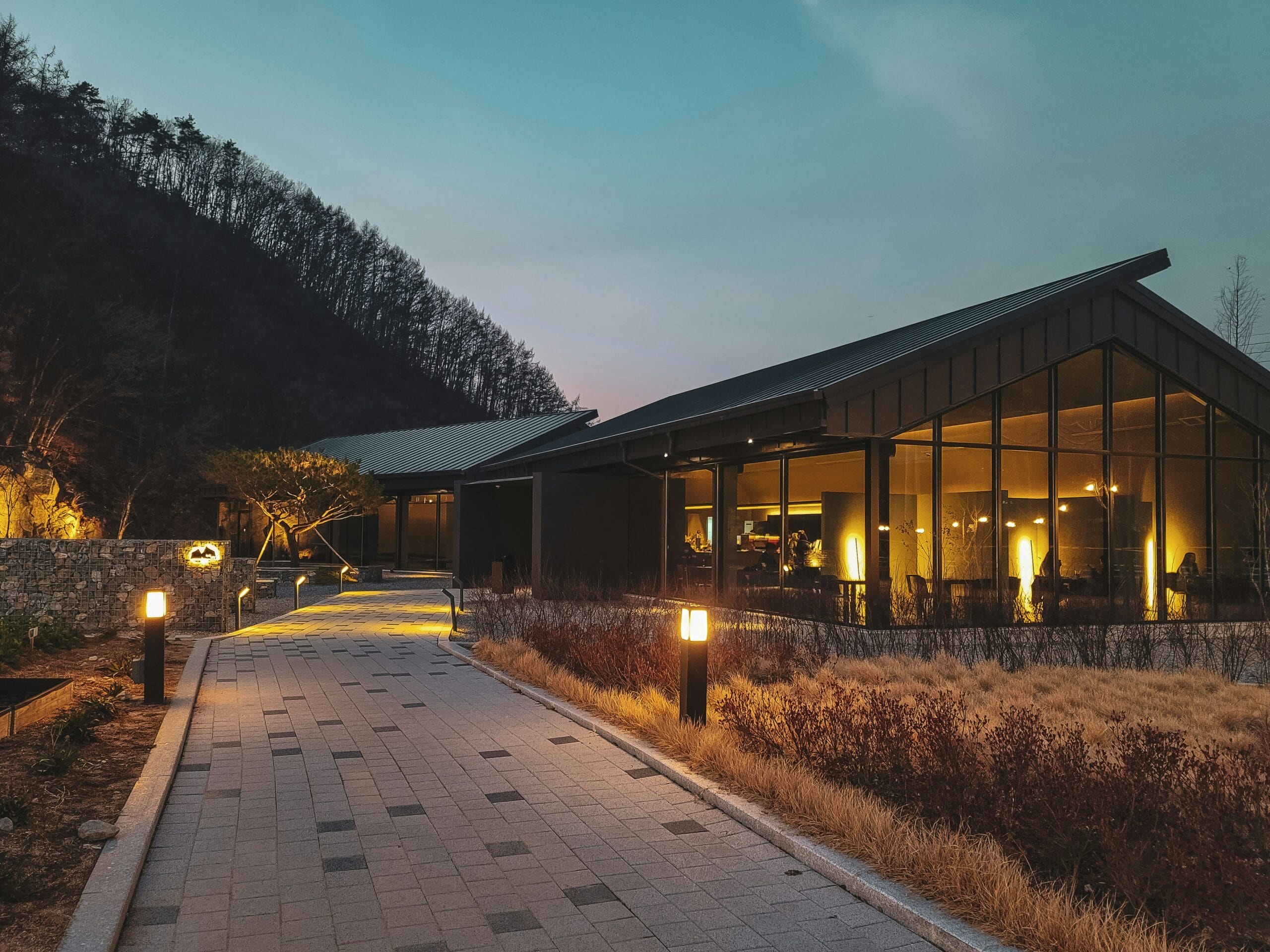 Modern building with large glass windows, surrounded by a pathway, trees, and soft evening lighting against a twilight sky.